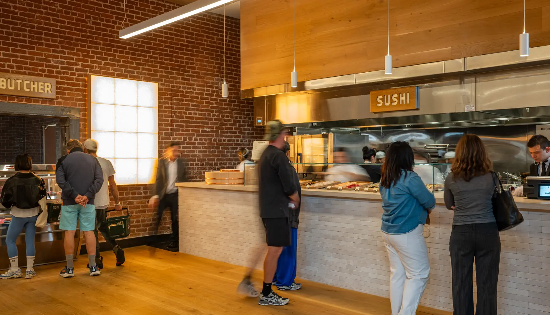 Shoppers at the butcher and sushi counters against a brick wall